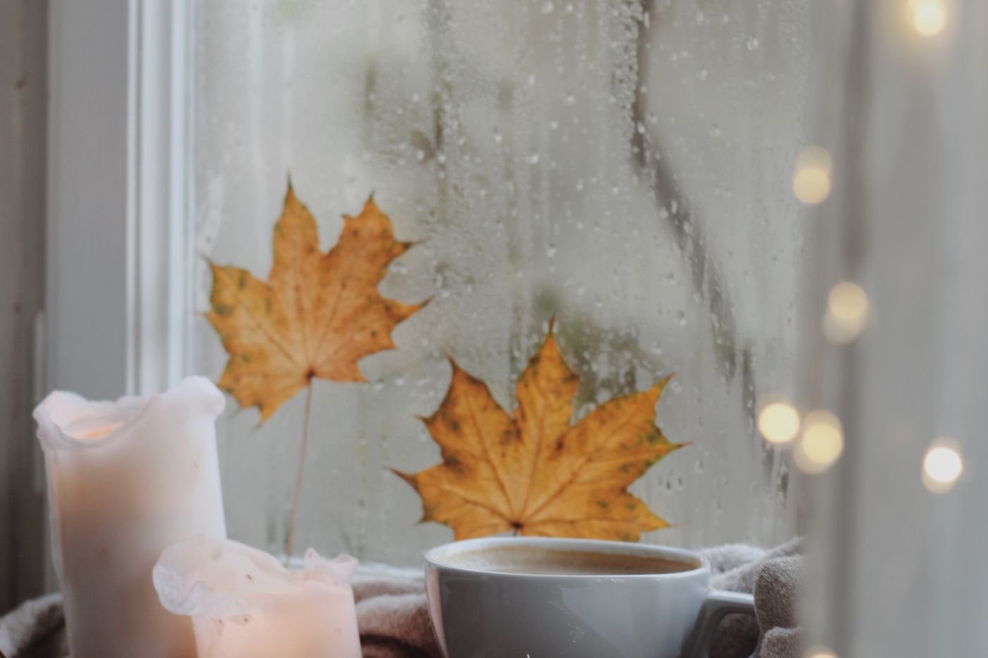 Regennasses Fenster mit Herbstblättern, davor Kerzen und eine Tasse Kaffee